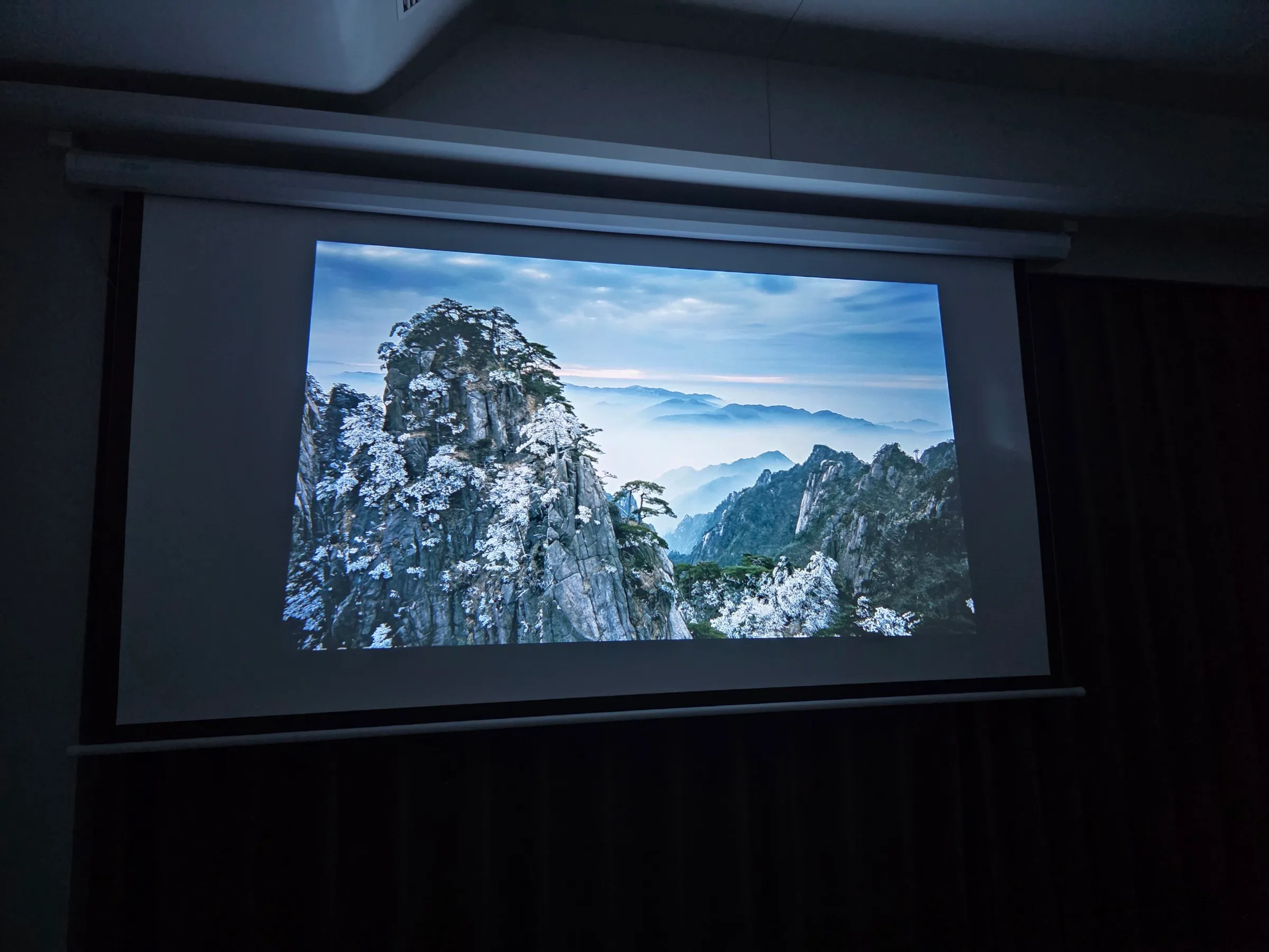 Ausgefahrene Projektionsleinwand mit Beamer-Bild einer Berglandschaft im abgedunkelten Wohnraum